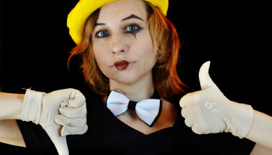 A young woman dressed as Columbine with a yellow beret, white bow tie and cream gloves. One hand is thumb up, the other thumb down